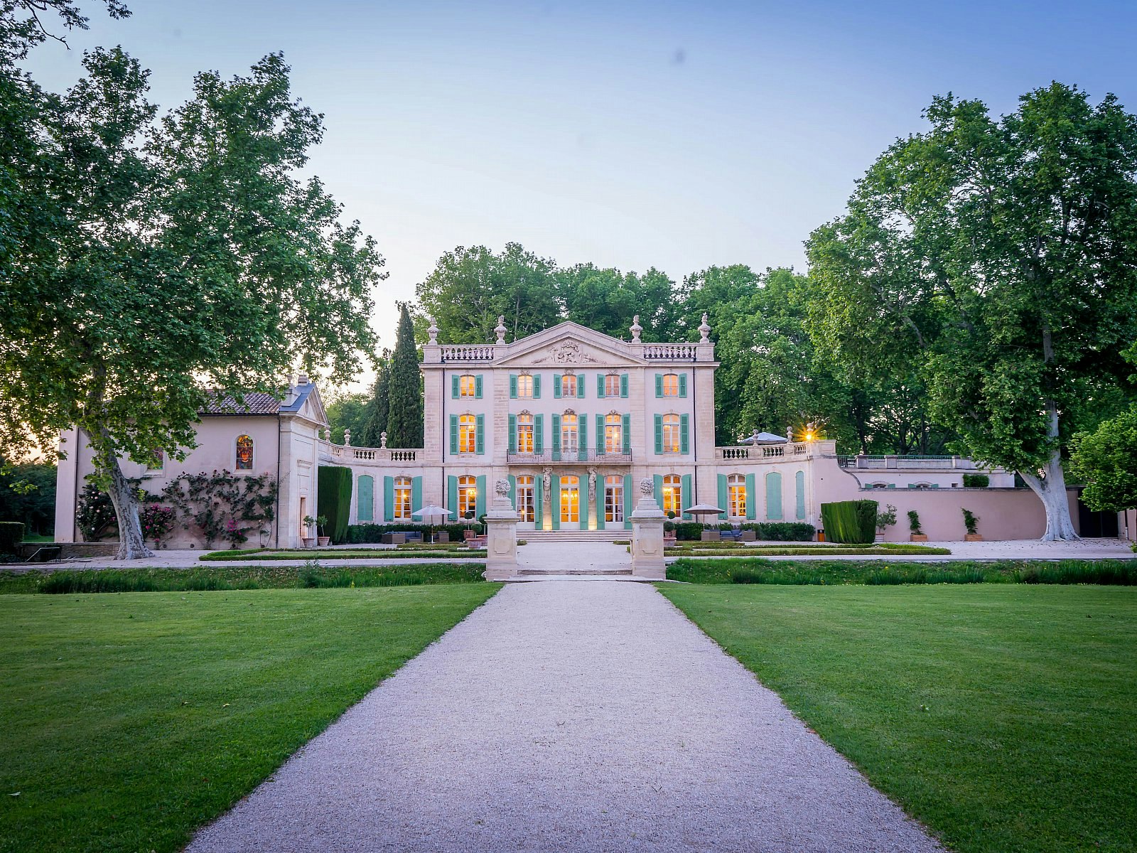 A gravel driveway leading up to an imposing stone French chateau, which has bright turquoise shutters on its windows; the building is surrounded by verdant greenery.